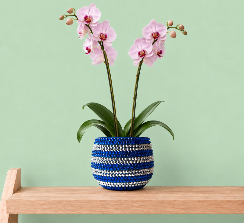 A potted artificial orchid with light pink flowers and green leaves, displayed in a blue and silver ceramic planter decorated with blue and silver crystals on a light wood shelf against a pale green background.