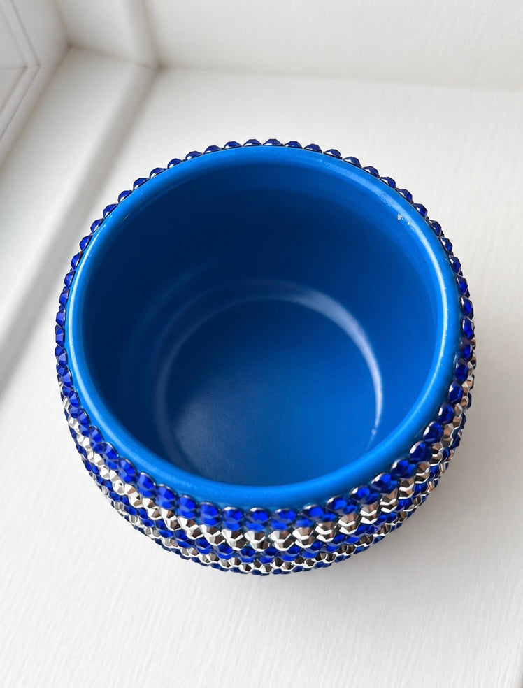 A small round ceramic plant pot decorated with rows of blue and silver crystals shown from a top-down angle on a white textured surface.