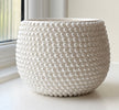 A round white ceramic planter with a textured beaded bubble pattern, sitting on a windowsill with natural light.