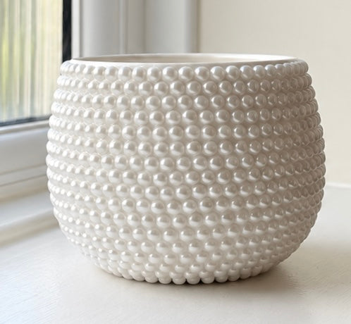 A round white ceramic planter with a textured beaded bubble pattern, sitting on a windowsill with natural light.